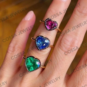 Puede incluir: Tres anillos con piedras preciosas en forma de corazón en rosa, azul y verde. Cada anillo tiene una banda dorada y un borde oscuro. Los anillos se muestran en los dedos de una persona.
