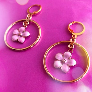 Puede incluir: Un par de pendientes de aro dorados. Cada pendiente presenta un delicado colgante de flor rosa claro suspendido dentro de un marco circular transparente. Sobre un fondo rosa vibrante.