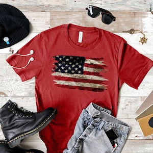 May include: Red t-shirt featuring a distressed American flag design. The shirt is paired with blue jeans, black boots, a black beanie, sunglasses, and other accessories. The shirt is a crew neck style.