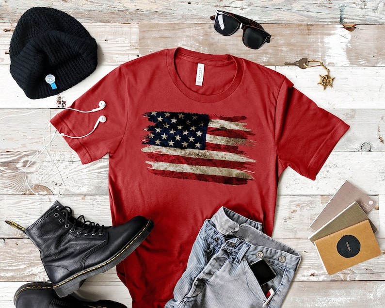 May include: Red t-shirt featuring a distressed American flag design. The shirt is paired with blue jeans, black boots, a black beanie, sunglasses, and other accessories. The shirt is a crew neck style.