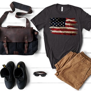 May include: A dark gray t-shirt with a distressed American flag design, paired with brown pants, black boots, a brown and black leather messenger bag, and sunglasses. The items are arranged on a white wooden surface.