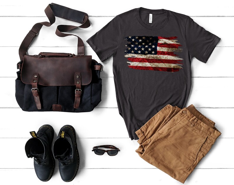 May include: A dark gray t-shirt with a distressed American flag design, paired with brown pants, black boots, a brown and black leather messenger bag, and sunglasses. The items are arranged on a white wooden surface.