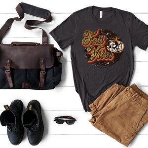 May include: A dark gray t-shirt with a graphic of a pumpkin and the text "Fall Ya'll" in a retro font. The graphic is surrounded by a leopard print pattern and a distressed circle.