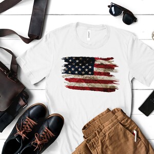 May include: White t-shirt featuring a distressed American flag design. The flag is rendered in red, white, and blue brushstrokes, with visible stars and stripes. The shirt is displayed with accessories like a leather bag, sunglasses, and a smartphone.