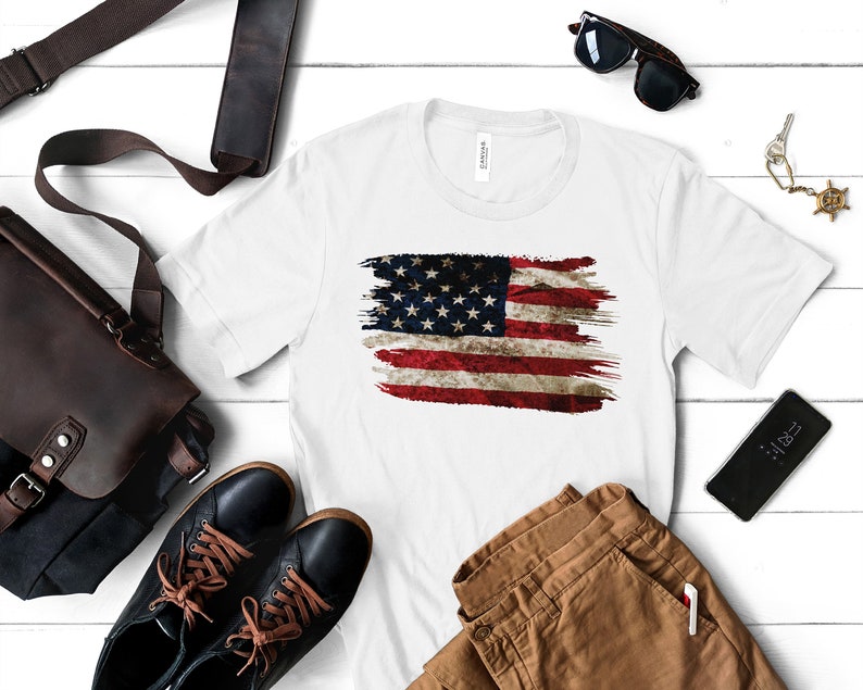 May include: White t-shirt featuring a distressed American flag design. The flag is rendered in red, white, and blue brushstrokes, with visible stars and stripes. The shirt is displayed with accessories like a leather bag, sunglasses, and a smartphone.