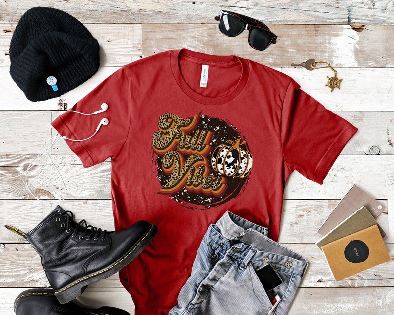 May include: Red t-shirt with a graphic design of the words "Fall Vibes" in a retro font with a leopard print pattern and a pumpkin with a cow print pattern.
