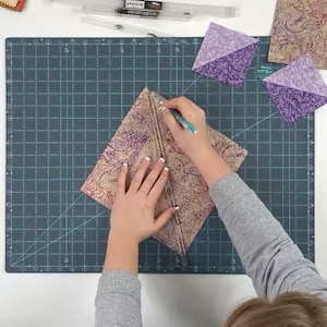 May include: A person using a ruler and pen to cut a patterned fabric on a dark green cutting mat. The fabric is tan with a purple floral design. Several purple fabric squares are also on the mat.