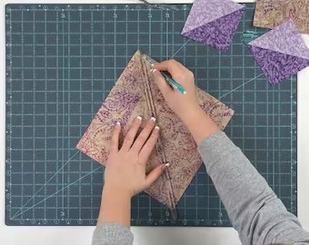 May include: A person using a ruler and pen to cut a patterned fabric on a dark green cutting mat. The fabric is tan with a purple floral design. Several purple fabric squares are also on the mat.