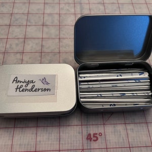 May include: A silver tin with a label that says "Amiya Henderson" and a butterfly design. The tin is open and contains a stack of silver metal strips.