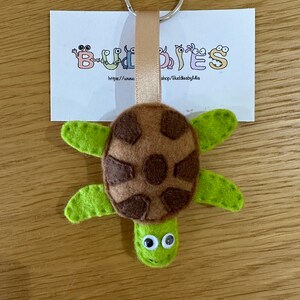 May include: A brown and green felt turtle keychain with googly eyes. The keychain is attached to a silver key ring and a brown ribbon. The keychain is on a wooden surface with a white card that says "Buddies" and a website address.