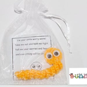 Pode incluir: Um verme de preocupação de crochê amarelo com olhos saltitantes em uma bolsa de organza branca. A bolsa tem um cartão com o texto "Eu sou seu pequeno verme de preocupação! Tire-me e segure-me forte. Diga-me suas preocupações e medos e tudo ficará bem."