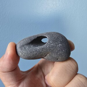 May include: A gray, smooth stone with a natural hole through the center. The stone is held in a hand against a light blue background. The stone's shape is rounded with a hole that appears to be naturally formed.