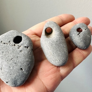 Könnte beinhalten: Drei glatte, graue Steine mit durchgebohrten Löchern. Die Steine sind unterschiedlich groß und werden in der Hand einer Person gehalten.