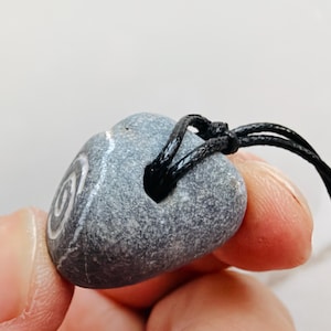 May include: A gray, heart-shaped stone pendant featuring a white spiral design etched into the surface. The stone has a smooth, speckled texture and is suspended from a thin, black cord that passes through a hole drilled near the top edge.