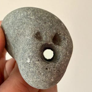 May include: A gray, weathered stone with a hole through the center and two smaller indentations above it. The stone has a smooth, rounded shape and a natural, textured surface. The stone is held in a hand.