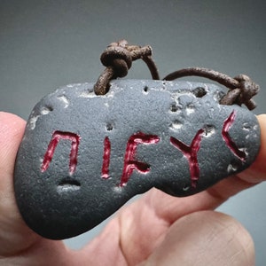 May include: A dark gray stone pendant with a red inscription that reads "NIY" hanging from a brown leather cord.