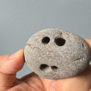 May include: A gray stone with three holes drilled into it, resembling a face. The stone is being held in a hand.