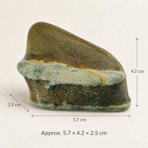Op de afbeelding: Een gladde, afgeronde steen met een donkerbruin bovengedeelte en een lichtere, lichtgroene band die horizontaal door het midden loopt. De steen meet zoals aangegeven ongeveer 5,7 cm in lengte, 4,2 cm in hoogte en 2,5 cm in diepte.