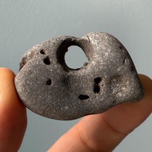 May include: A gray, weathered stone with a large, circular hole in the center and several smaller holes. The stone is held between fingers, showcasing its unique shape and texture. This natural stone could be used for jewelry or home decor.