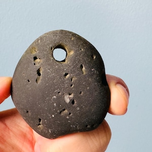 Könnte beinhalten: Ein dunkelgrauer, glatter, runder Stein mit einem durchbohrten Loch in der Mitte. Der Stein hat ein natürliches, verwittertes Aussehen mit kleinen Vertiefungen und Gruben.