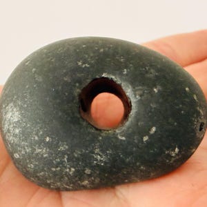 Könnte beinhalten: Ein dunkelgrauer, abgerundeter Stein mit einem Loch in der Mitte. Der Stein hat eine glatte, abgenutzte Textur und wird in der Hand einer Person gehalten. Das Loch ist kreisförmig und scheint poliert zu sein. Der Stein hat eine natürliche, organische Form.