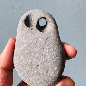 Puede incluir: Una piedra lisa de color gris con dos agujeros tallados, que se asemejan a los ojos de un búho. La piedra está sostenida en una mano.