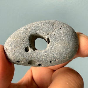 May include: A gray, oval-shaped stone with a large, circular hole in the center. The stone has a rough, textured surface and several smaller holes. The stone is being held up by a person's fingers.