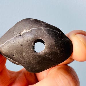 May include: A smooth, black stone with a hole drilled through the center. The stone has a natural, rough texture and a white line running through it.
