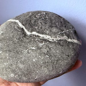 May include: A smooth, gray and white stone with a white vein running through it. The stone is round and has a rough texture.
