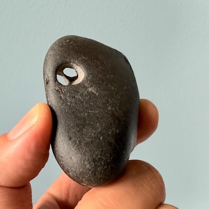 May include: A dark gray, smooth stone with a natural hole, held between fingers. The stone's surface is slightly textured, and the hole is circular. The background is a light blue.