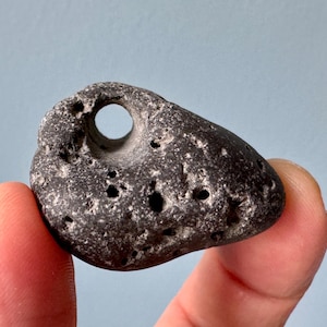 May include: A dark gray, smooth, round stone with a hole through the center. The stone has a rough, textured surface and is covered in small, white dots.