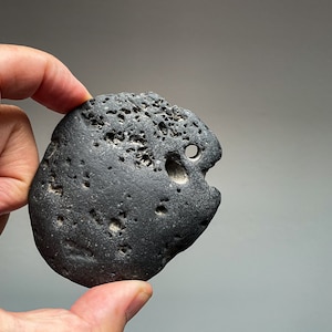 Könnte beinhalten: Ein dunkelgrauer, glatter, runder Stein mit einem Loch in der Mitte. Der Stein hat eine raue, strukturierte Oberfläche und ist mit kleinen schwarzen Punkten bedeckt.