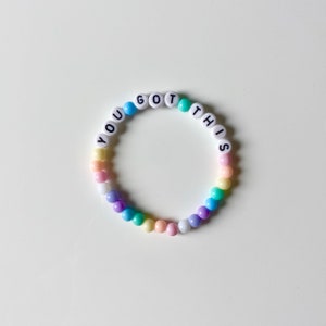 May include: A pastel rainbow beaded bracelet with the words "YOU GOT THIS" spelled out in white letters.