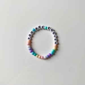 May include: A pastel-colored beaded bracelet with the words "YOU ARE ENOUGH" spelled out in white letters.