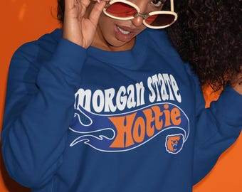 May include: A blue sweatshirt with the words "Morgan State Hottie" in white and orange lettering. The sweatshirt has a retro design and a small bear logo. The background is orange.