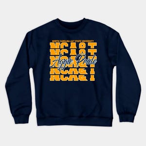May include: A navy blue long-sleeve sweatshirt featuring the text NORTH CAROLINA A&T STATE UNIVERSITY in yellow, followed by five stacked rows of the yellow text NCA&T, and the white cursive script text Aggie Pride layered across the center.