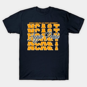 May include: A navy blue short-sleeve t-shirt featuring the text North Carolina A&T State University in orange at the top, followed by five rows of the orange text NC A&T, and the white cursive script text Aggie Pride layered across the center.