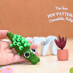 May include: A green crocheted crocodile fidget toy with large black eyes and green bead accents. The text "No Sew PDF PATTERN Crocodile Fidget" is visible in the background. A small potted plant with red leaves is also in the background.