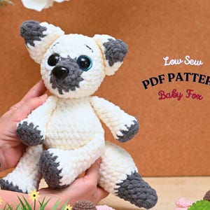 May include: A crocheted white and gray baby fox plush toy with big blue eyes. The toy is being held by a hand. The text "Low Sew PDF PATTERN Baby Fox" is in the background.