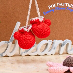 Puede incluir: Llavero de guantes de boxeo de crochet rojo colgando de una cuerda blanca. Los guantes están colgando sobre la palabra "dream" en letras blancas. El fondo es una superficie marrón texturizada.