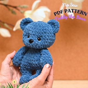 Peut inclure: Un ours en peluche bleu crochet&eacute; avec des yeux noirs et un nez noir. L'ours est tenu dans les mains d'une personne. Le texte "No Sew PDF PATTERN Teddy the Bear" est visible en arri&egrave;re-plan.
