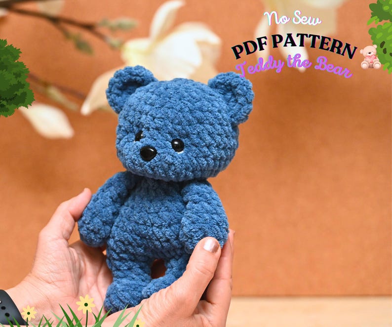 Peut inclure: Un ours en peluche bleu crochet&eacute; avec des yeux noirs et un nez noir. L'ours est tenu dans les mains d'une personne. Le texte "No Sew PDF PATTERN Teddy the Bear" est visible en arri&egrave;re-plan.