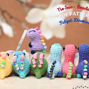 Può includere: Una collezione di sei piccole figure di dinosauri all'uncinetto in giallo, verde acqua, verde, azzurro, rosa e viola. Ogni dinosauro presenta una fila di perline di plastica colorate sul dorso. Il testo "No Sew - Beaded PDF PATTERN Fidget Dinosaur" è visibile.