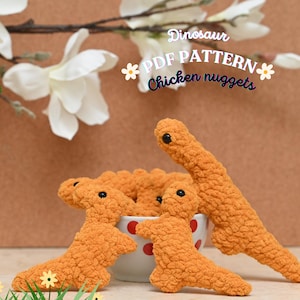 Puede incluir: Se muestran nuggets de pollo de dinosaurio de ganchillo naranja con ojos negros. La imagen incluye el texto "Dinosaur PDF Pattern Chicken nuggets". Están en un cuenco blanco con lunares rojos. El fondo tiene flores blancas y una superficie marrón.