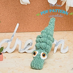 May include: A green crocheted crocodile toy with large eyes. The toy is sitting on a white sign that says "dream". The sign is on a brown corkboard background.