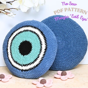 Könnte beinhalten: Zwei gehäkelte Kissen, eines mit einem blauen und türkisfarbenen bösen Auge und das andere einfarbig blau. Die Kissen liegen auf einer gefliesten Oberfläche mit rosa Blumen. Der Text "No Sew PDF PATTERN Magic 'Evil Eye'" befindet sich in der oberen rechten Ecke des Bildes.