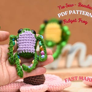 May include: A handmade green and purple crochet frog with beaded accents. The frog has black eyes and is held in a hand. The text "No Sew - Beaded PDF Pattern Fidget Frog" is visible in the image.
