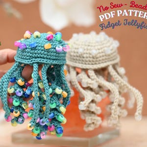 May include: Two crocheted jellyfish toys with dangling tentacles adorned with colorful plastic beads. The teal jellyfish features multi-colored beads, while the beige one has clear beads. Text in the corner reads: No Sew - Beaded PDF PATTERN Fidget Jellyfish.