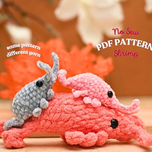 May include: Three crocheted shrimp toys in shades of pink and gray. The largest shrimp is coral-colored, with two smaller shrimp in gray and pink resting on top. The image includes the text "No Sew PDF Pattern Shrimp" and "same pattern different yarn."