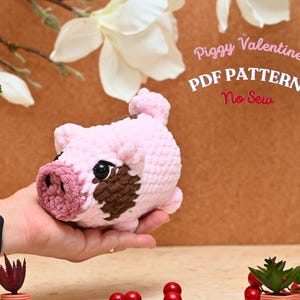 Può includere: Un giocattolo di maiale amigurumi in crochet rosa e marrone. Il giocattolo è nel palmo di una mano. Il testo "Piggy Valentine PDF PATTERN No Sew" è sullo sfondo.